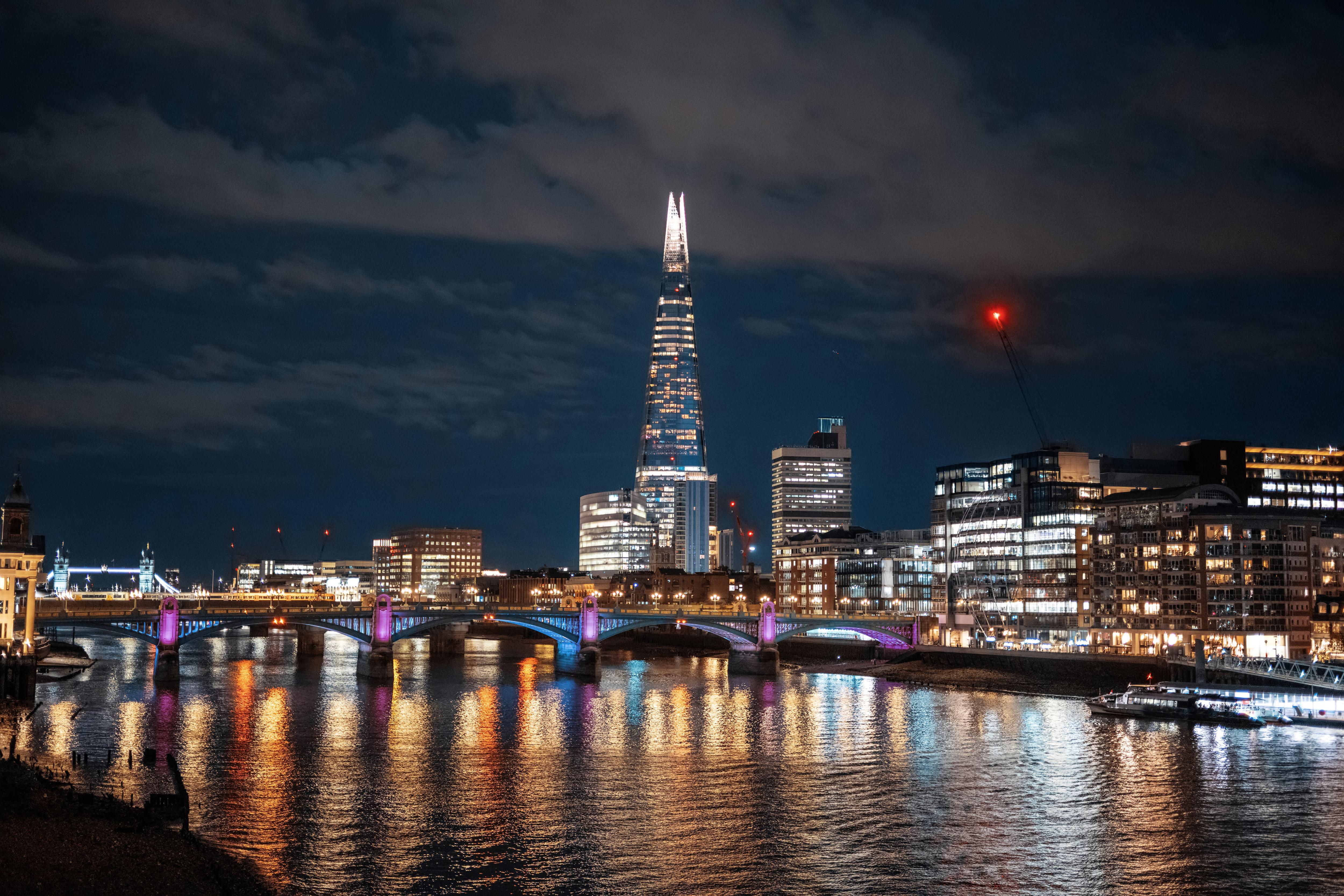 A city skyline at night with tall skyscrapers illuminated against a dark sky, reflecting on the water of a nearby lake or river. The scene captures the essence of a bustling metropolis with bright lights and urban architecture.