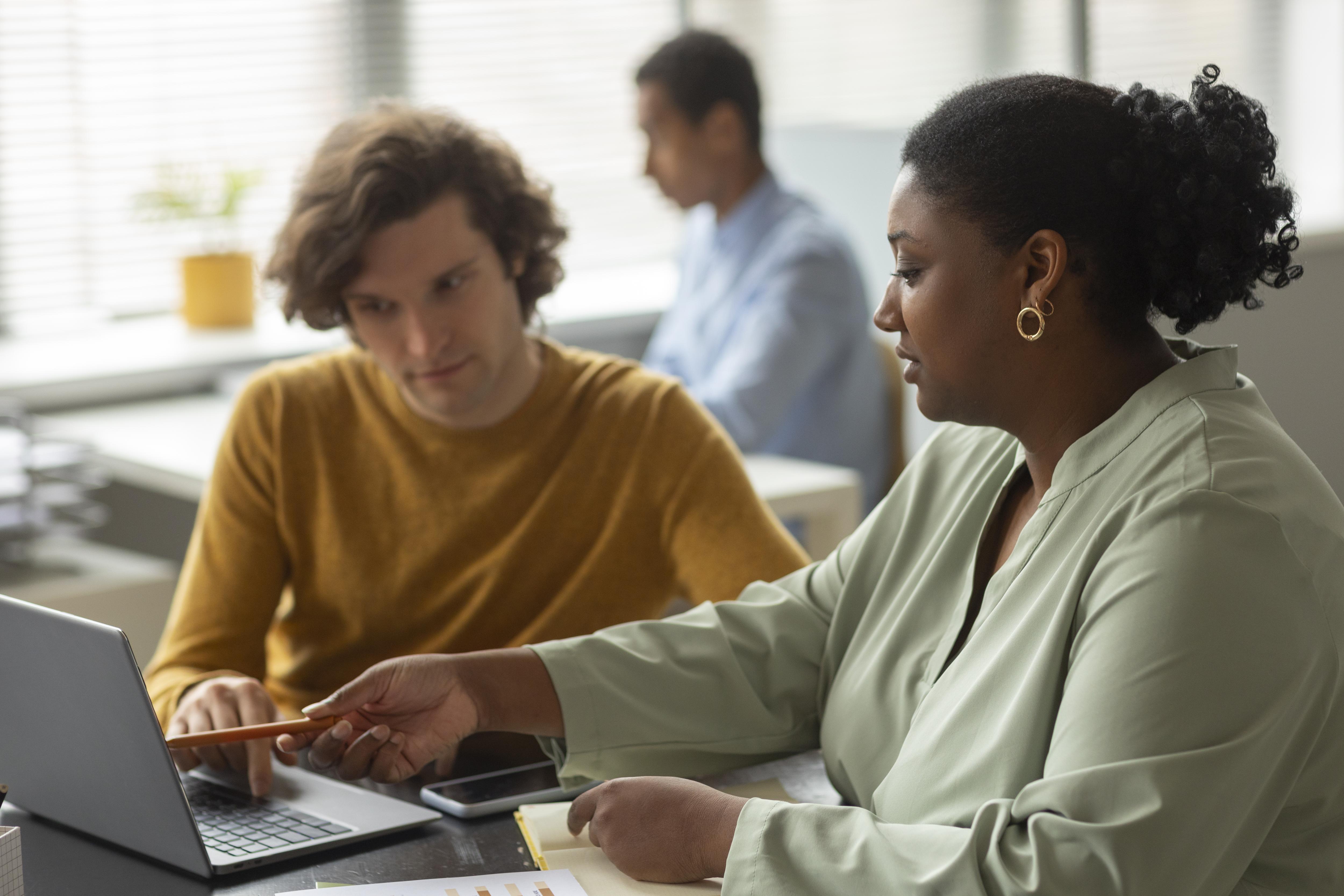 A woman and a man are seated indoors, both looking at a laptop. The setting appears to be an office building, with a wall in the background. They are engaged in what seems to be work-related activities, focusing on the computer together.