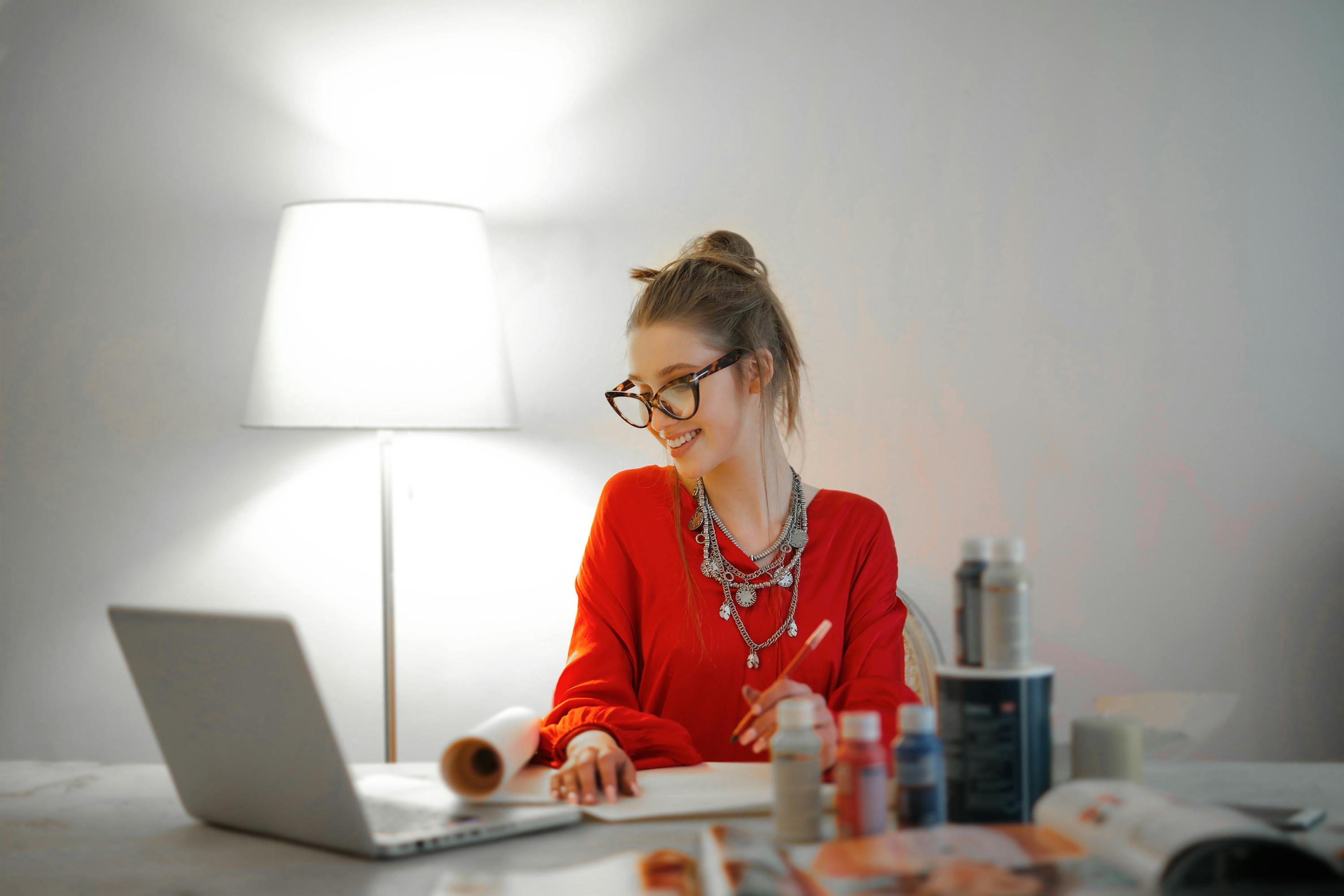 A woman with glasses and a red top smiles whilst working on her laptop at her desk.