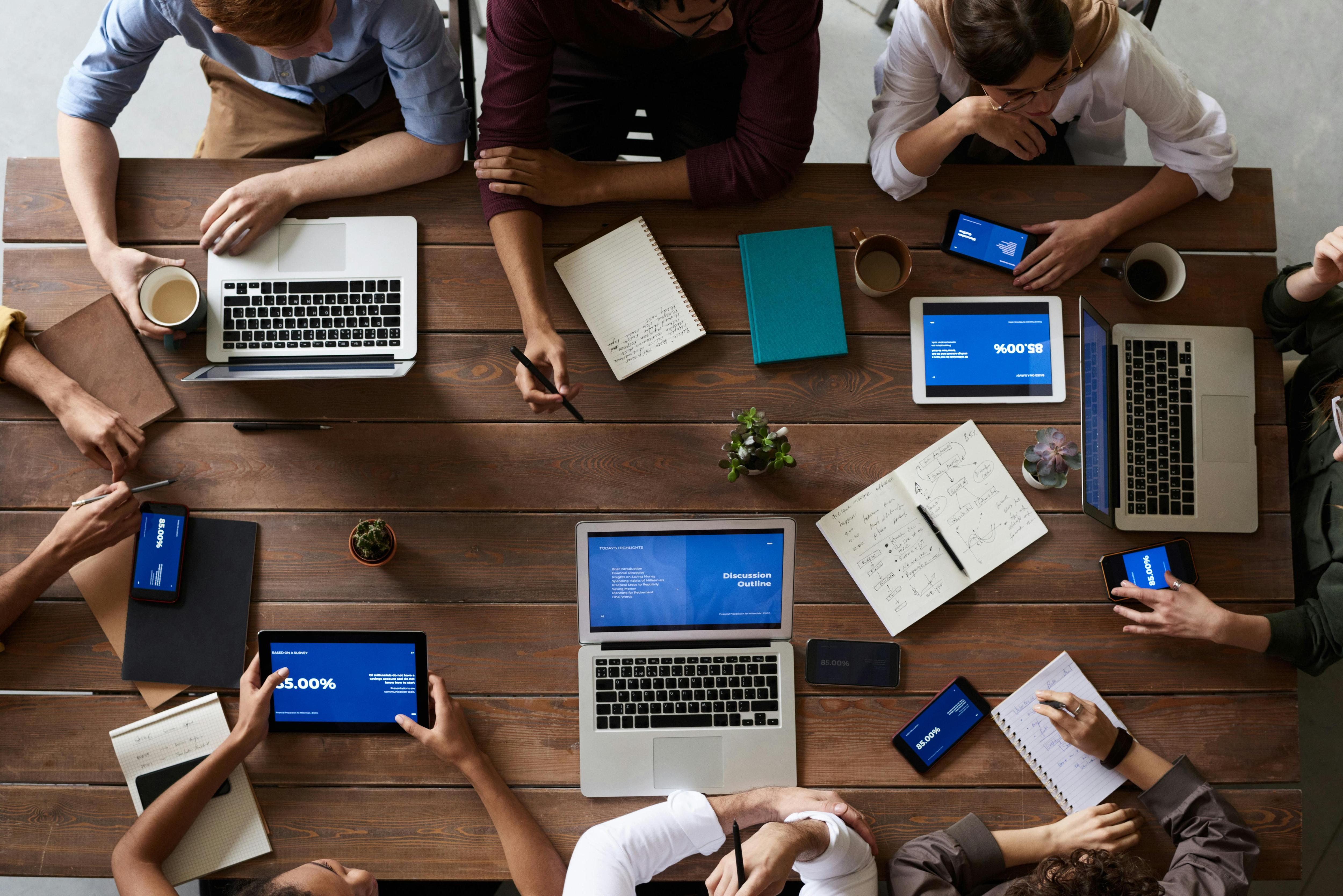 A birds eye image of a group of people sitting at a wooden desk with their notes and laptops