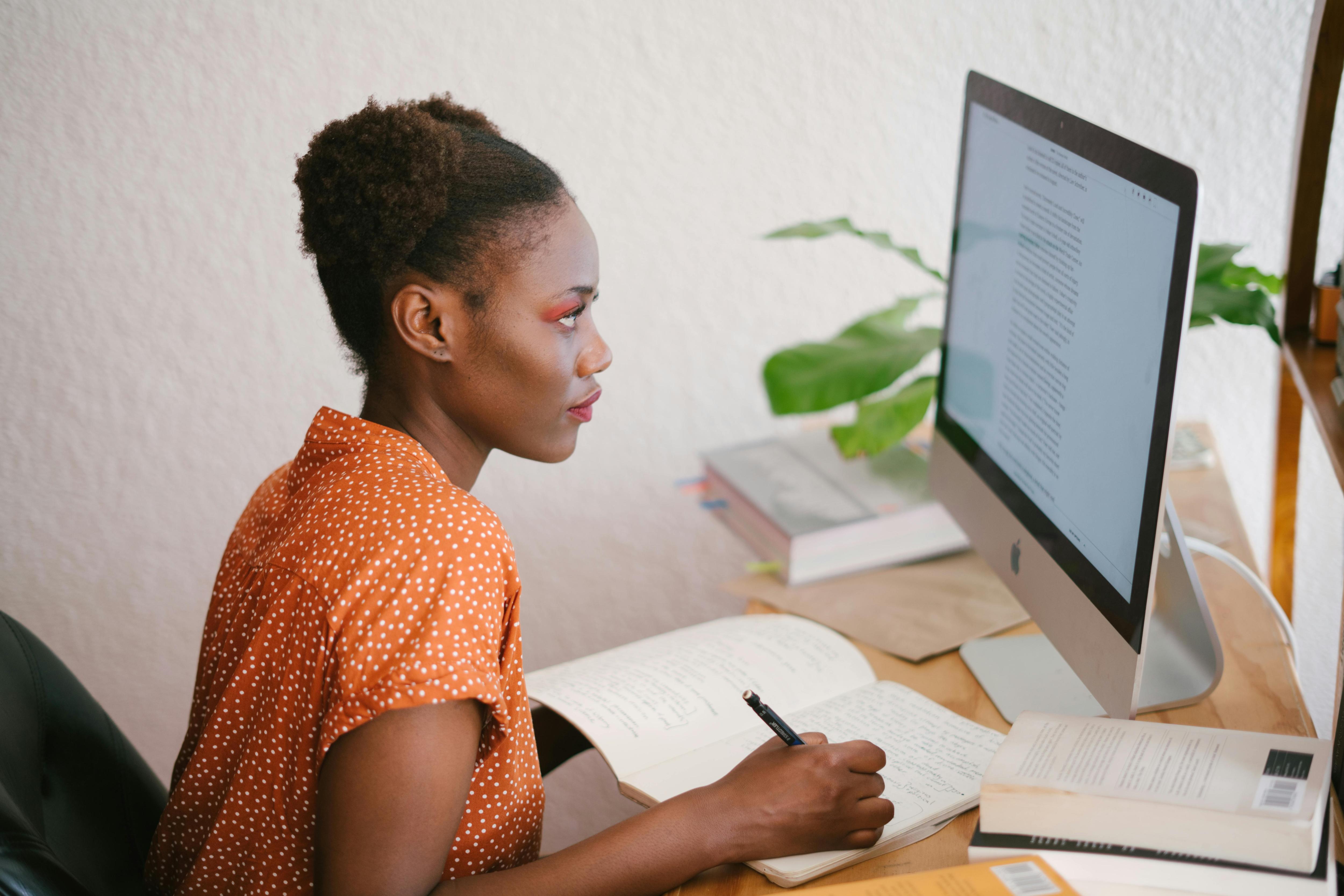 An image of a person sitting at her desk taking notes while looking at her computer screen